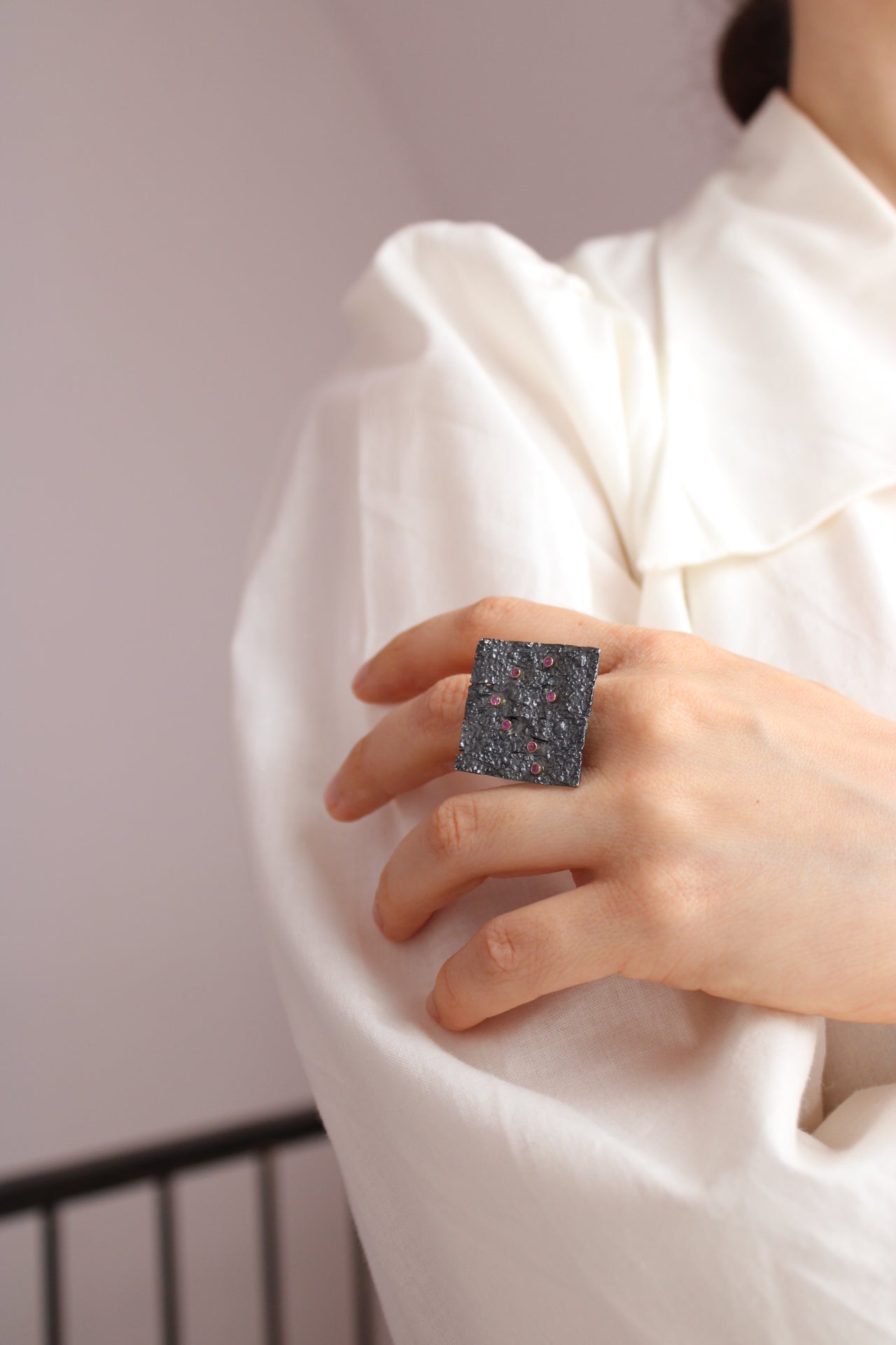 A close-up of a person's hand wearing a large square ring with a dark, textured, speckled surface, set against a white draped garment.