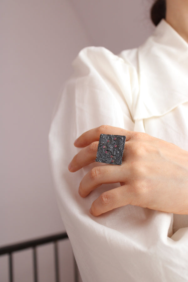 A close-up of a person's hand wearing a large square ring with a dark, textured, speckled surface, set against a white draped garment.