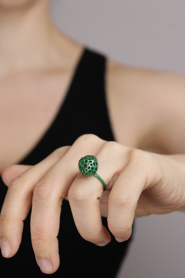 Hand wearing a green ring with an embedded ruby on a neutral background